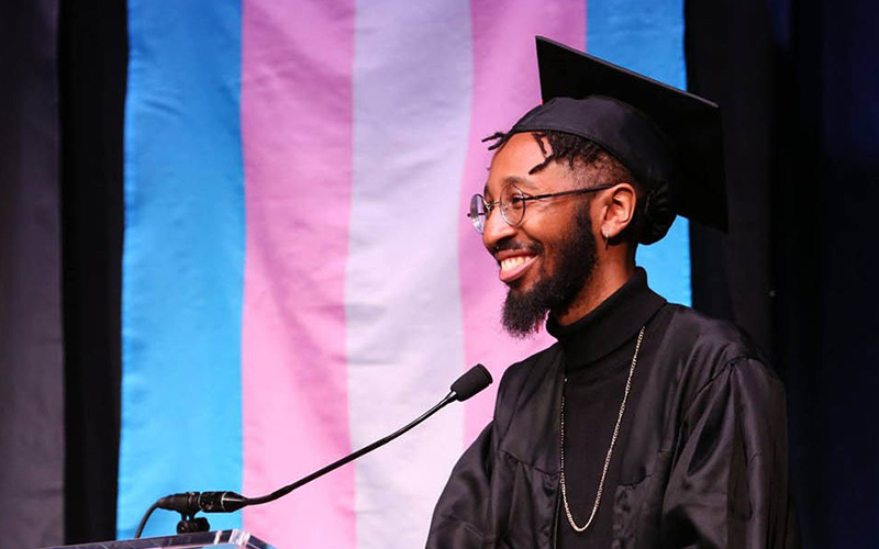 A student in a graduation cap and gown smiles in front of a trans flag