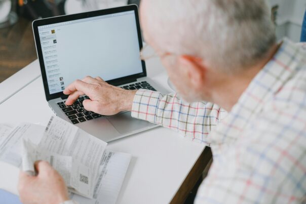 A man types on a laptop while also holding receipts and other financial documents.