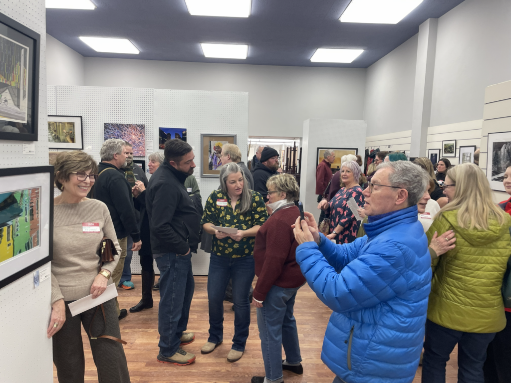 Attendees gather inside an art gallery during a reception, viewing framed artwork displayed on white walls and display panels. Several people are engaged in conversation throughout the room, while one attendee in a blue jacket photographs a piece of art with a smartphone. The gallery features wood flooring, bright overhead lighting and a variety of colorful photographs and paintings on display.
