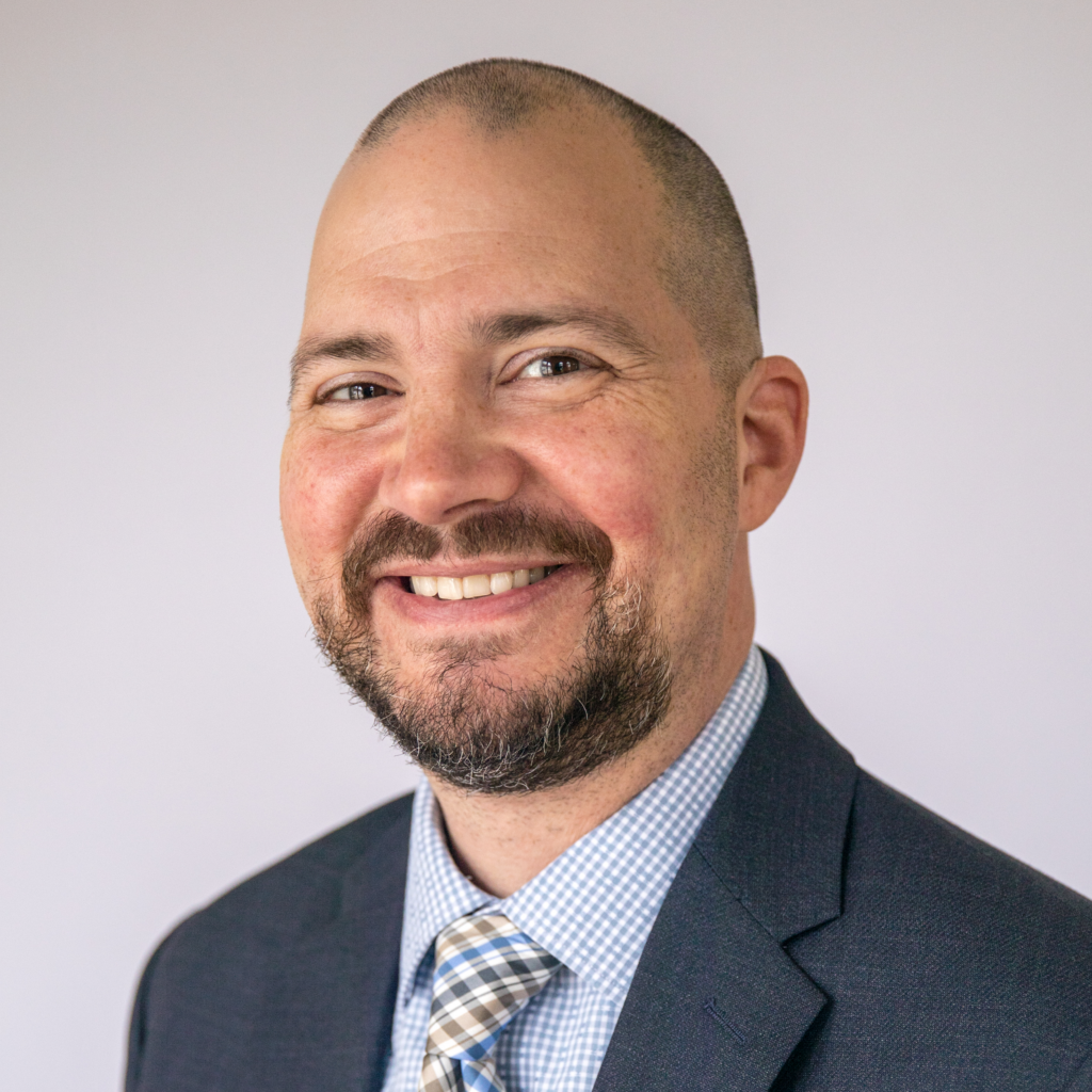 Professional headshot of a person wearing a dark suit jacket, light blue checkered dress shirt and patterned tie. The person has a closely shaved head and neatly trimmed beard and is photographed against a light gray background. The image is cropped from the chest up and is evenly lit.