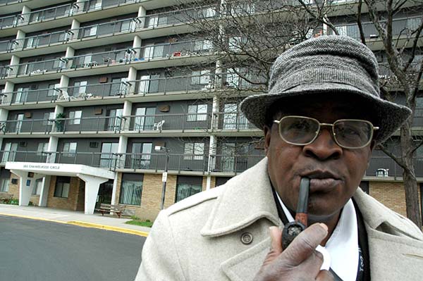 Alfred McMoore stands in front of apartment building with a pipe in his mouth