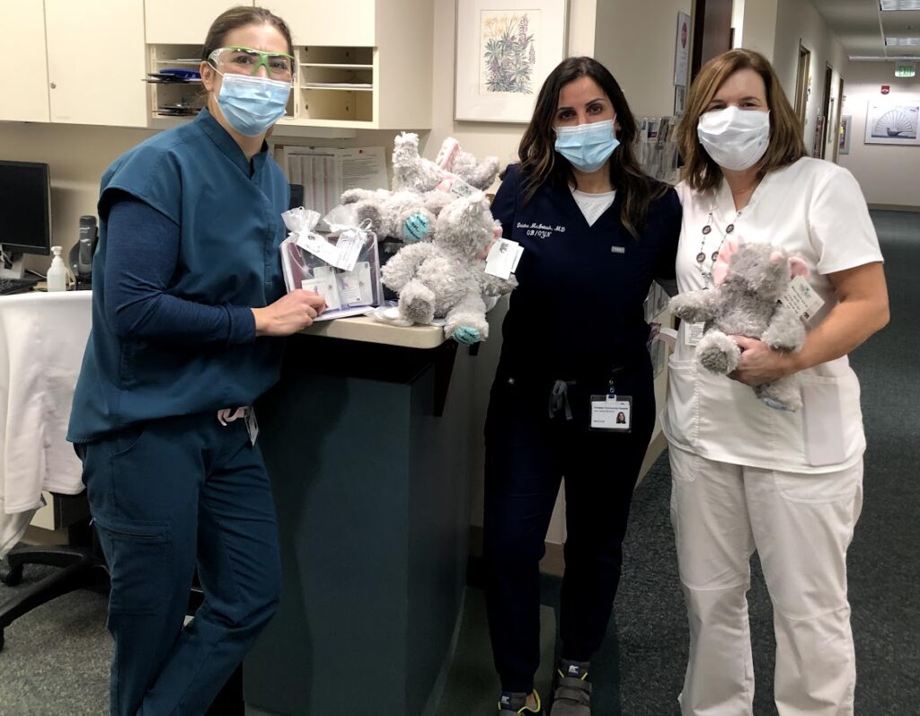 Three masked people pose in a hospital hallway next to stuffed elephants as part of the Forget-Me-Not Baskets program.