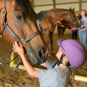 A young girl wearing a purple helmet affectionately greets a horse in a barn at Victory Gallop.
