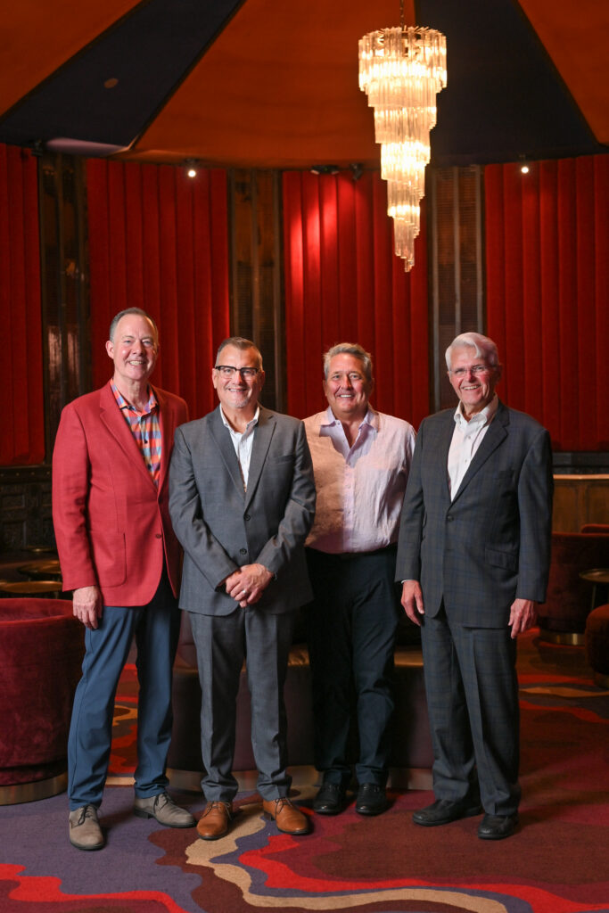 Four people stand together in an elegant lounge with deep red walls, plush seating and a large tiered chandelier hanging overhead. One person wears a coral blazer, two wear gray suits and one wears a light-colored dress shirt with dark trousers. The group is positioned beneath the chandelier on a patterned carpet, with warm lighting highlighting the sophisticated interior setting.