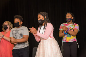 Three children and one adult, all wearing masks, applaud a dancer's performance