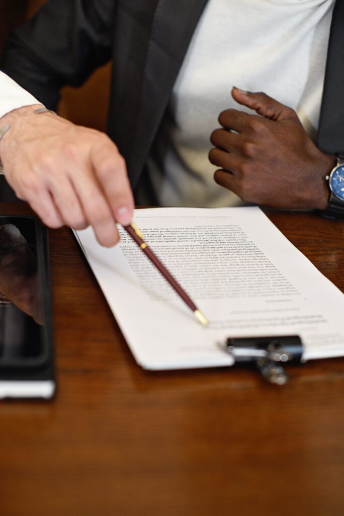 Two people seated at a wooden table reviewing a document on a clipboard. One person holds a pencil and gestures toward the paperwork, while another person wearing a wristwatch sits across the table. A smartphone is partially visible beside the document, suggesting a professional meeting or financial discussion.