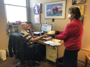 A woman wearing a medical mask hands a package to a man seated on the desk, while he talks on the telephone