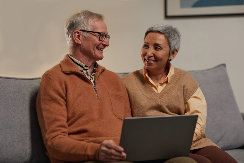 An older couple smiles at one another on a couch, while holding a laptop