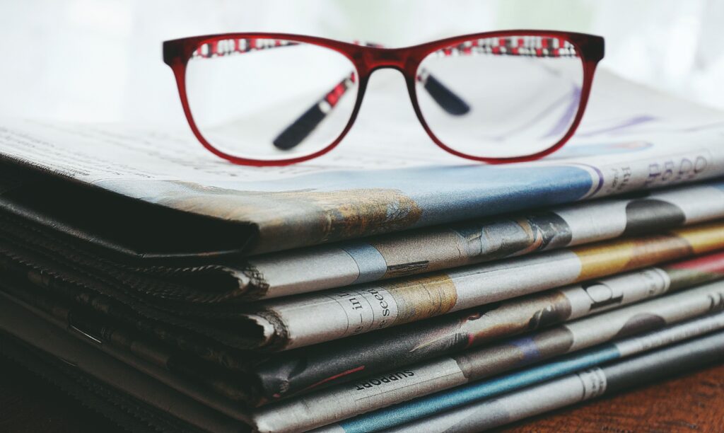 A pair of red-framed eyeglasses resting on top of a stack of folded newspapers. The image is a close-up with the glasses in sharp focus and the newspapers slightly blurred, creating a professional reading and research theme. The newspapers are stacked on a wooden surface, with visible printed text and muted tones in the background.