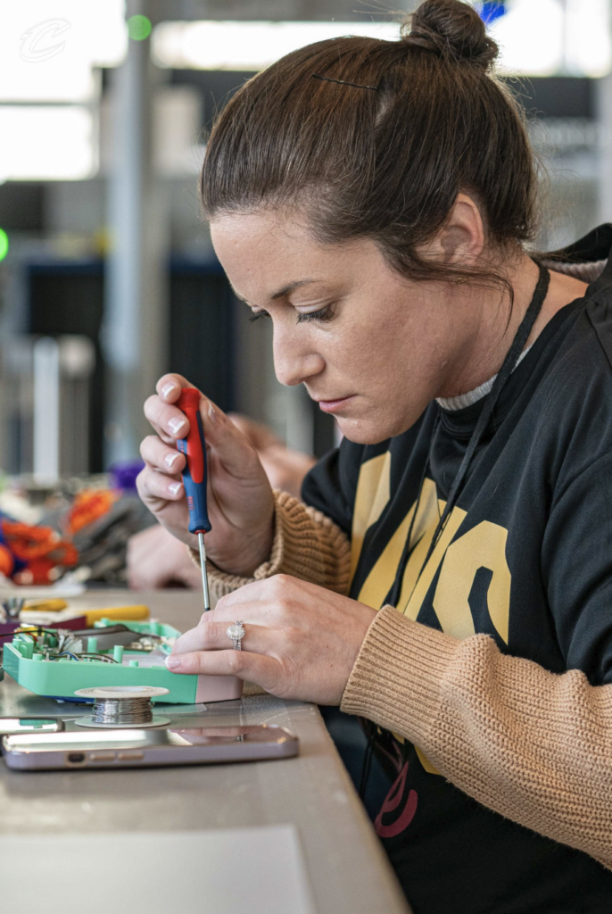 A woman works on an electronic device at a workshop table, using a screwdriver to make a repair. Small tools, wiring, and a spool of solder are arranged on the table near the device, while another person and additional equipment are visible in the background. The image shows a hands-on electronics repair activity in a well-lit indoor setting.