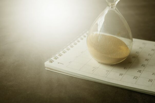 An hourglass filled with golden sand resting on an open monthly calendar planner. Soft natural light illuminates the scene, with the calendar dates partially visible beneath the glass. The image symbolizes deadlines, scheduling, time-sensitive planning, and year-end preparation.