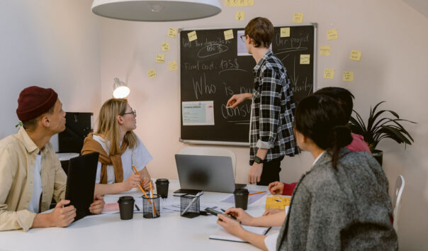 A group of people sit at a table at a meeting while a person stands at a chalkboard.