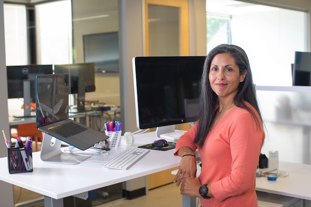 A business owner poses standing at a desk near a computer screen and a laptop computer. 