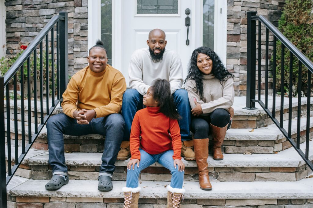 Four people sit on the stone steps of a house in front of a white entry door and stone exterior wall. One child in a red turtleneck sweater sits on the lower step, while three other people sit behind them. Black metal railings frame both sides of the staircase, and the group faces the camera outdoors in daylight.
