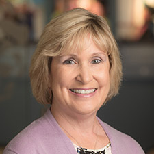Professional head-and-shoulders portrait of a person with short, light-colored hair, wearing a light purple cardigan over a patterned top and a necklace. The person is facing the camera with a smile. The background is softly blurred, creating a studio-style corporate headshot appearance.