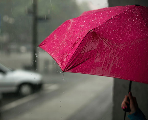 A red umbrella is being held in the rain along the side of the street, with parked cars in the distance. 