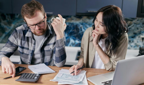 Two people are sitting at a table looking over paperwork and a calculator, confused.