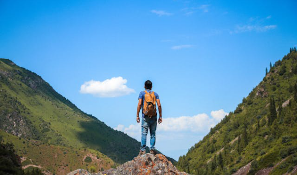 We see the back of a man as he stands at the top of a hill looking up at a mountainous landscape.