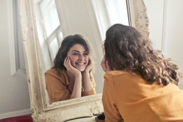 A girl looks into the mirror with a smile on her face, as she rests the bottom of her chin on her open hands.