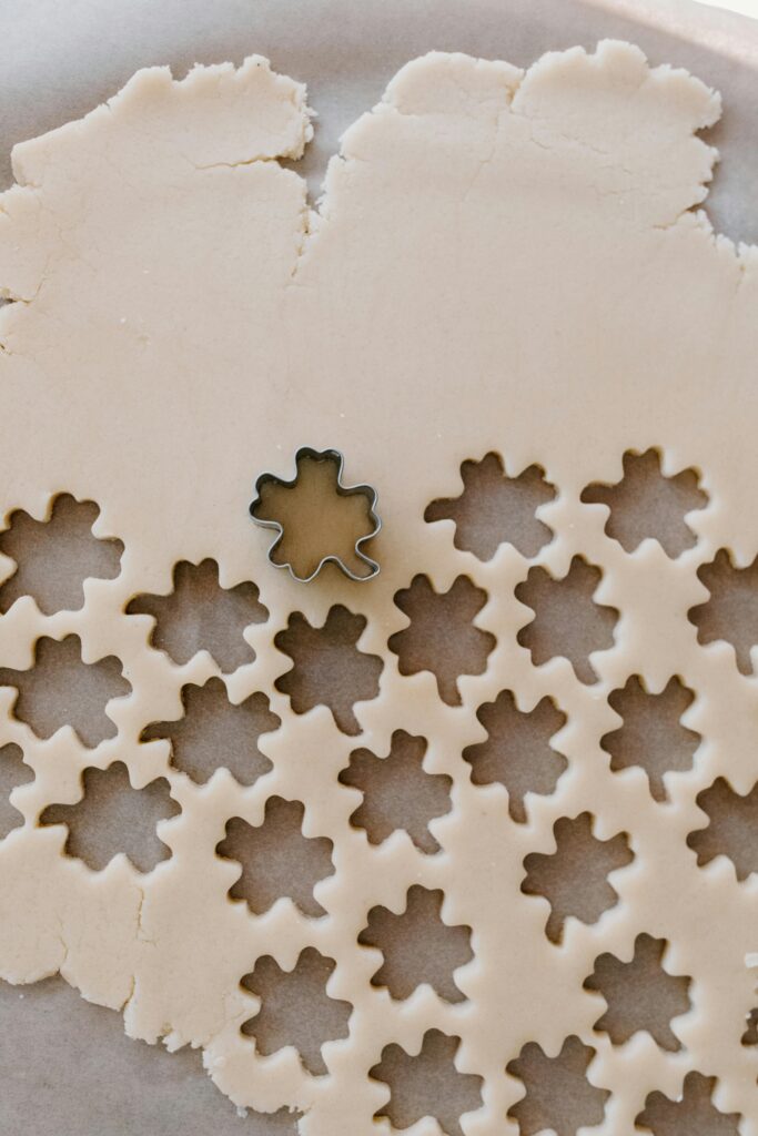 A cookie cutter is making cutouts of a leaf pattern on a flat sheet of cookie dough.