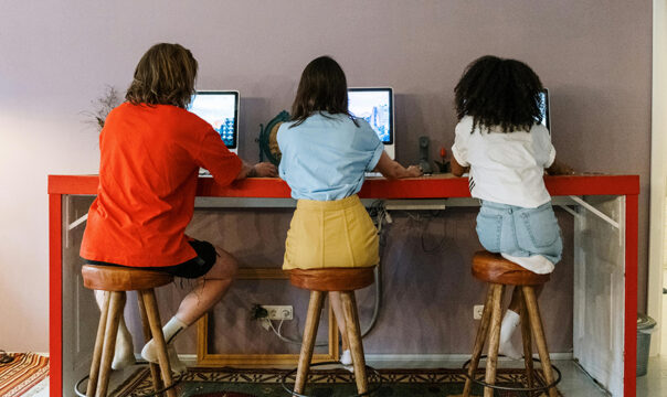 Three people sit on stools at a table, each working on a laptop, with their backs to the camera.
