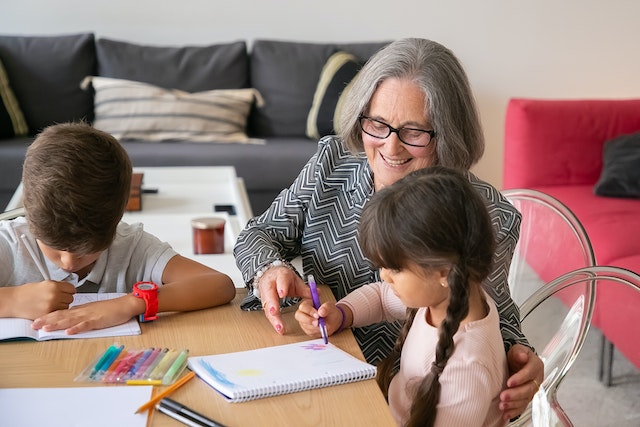 A woman is sitting at a table with what appear to be her grandchildren, who are drawing and coloring on notebook paper. 