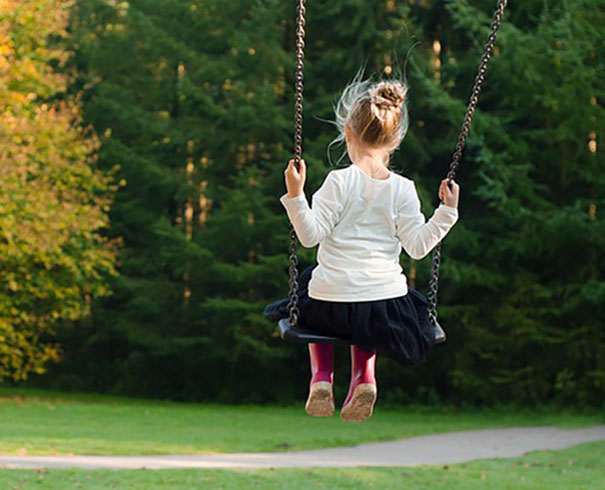 A girl holds onto the chains of a swing, while facing a row of trees.